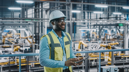 Happy African American Car Factory Engineer in High Visibility Vest Using Tablet Computer. Automotive Industrial Facility Working on Vehicle Production on Automated Technology Assembly Plant.