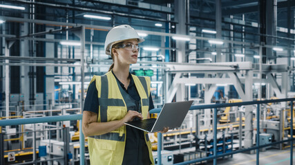 Portrait of Female Automotive Industry 4.0 Engineer in Safety Uniform Using Laptop at Car Factory Facility. Happy Assembly Plant Specialist Working on Manufacturing Modern Electric Vehicles.