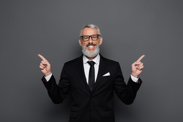 positive middle aged businessman in glasses pointing with fingers aside on grey
