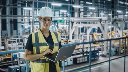Portrait of Female Automotive Industry 4.0 Engineer in Safety Uniform Using Laptop at Car Factory Facility. Confident Assembly Plant Specialist Working on Manufacturing Modern Electric Vehicles.