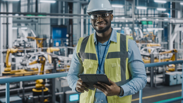 Portrait of Automotive Industry Engineer in Safety Uniform Using Tablet at Car Factory Facility. Happy Assembly Plant African American Man Specialist Working on Manufacturing Modern Electric Vehicles.
