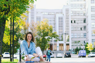 Fototapeta premium Urban portrait of young elegant business woman in eyeglasses and casual clothes sitting on a bench in the city. Drinking healthy smoothie, reading a book and working.