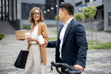 Fototapeta premium Asian businessman and caucasian businesswoman walking, drinking coffee and talking in city. Concept of business cooperation. Idea of freelance and remote work. Man with bicycle. Young woman with food