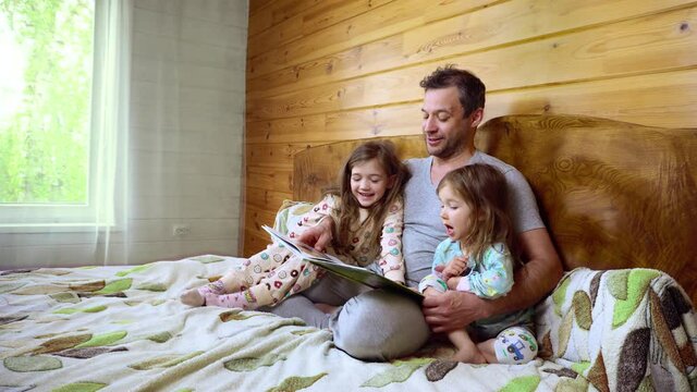 Father sits on the bed with his cute little daughtersin and reads the book. In the cozy bedroom. Girles in pajamas. In a weekend morning. Spending the weekend time. Indoors. . High quality 4k footage