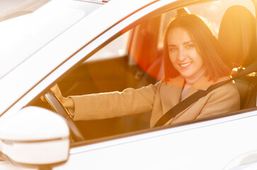 A young woman in coat is driving a car. Smiling female looks at camera and smiles in warm natural light. Taking driving courses