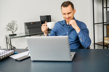 Male freelancer drinking coffee, using a laptop for work in a office. A millennial guy works on outsours, receiving emails, conducts correspondence with customer