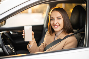 Young happy woman sits inside of new modern car, holds smartphone with empty blank screen, demonstrating new mobile app for drivers, copy space
