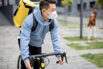 Courier in medical mask riding bicycle delivering food in city. Concept of shipping and logistics during Coronavirus pandemic. Idea of profession and job. Asian man wearing warm clothes