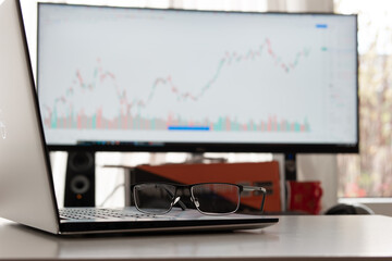 Clear reading glasses, black rim, on a laptop keypad. Blurred large computer monitor in the background showing stock market graph. No people
