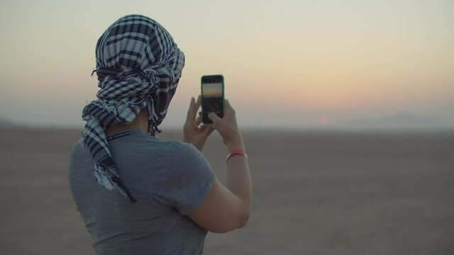 Woman in checkered keffiyeh taking picture of sunrise on mobile phone standing in the desert. Caucasian female tourist enjoy desert adventure.