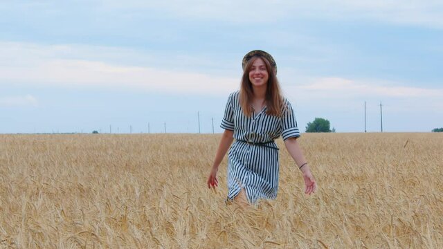 Happy brown-haired girl in dress goes across field, touches ears of wheat with hands in countryside. Woman farmer rejoices at good crop. Young lady walks on farmland, smiling in summer. Front view