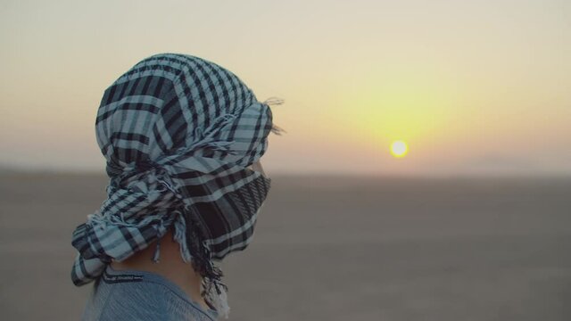 30s woman wearing black and white checkered keffiyeh standing in the desert looking at camera. Caucasian female tourist taking off headdress and smiling at camera. Desert sunrise.
