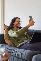 Vertical medium shot of young man in video call smiling to smartphone with laptop on the sofa