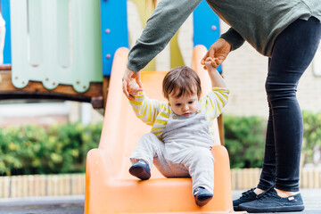 Middle-aged young mother woman in the park helping her 1 year old baby down the slide. real...