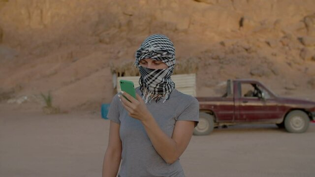 Woman wearing checkered keffiyeh standing in the desert using cell phone. Early morning in desert mountains. Using smartphone everywhere.