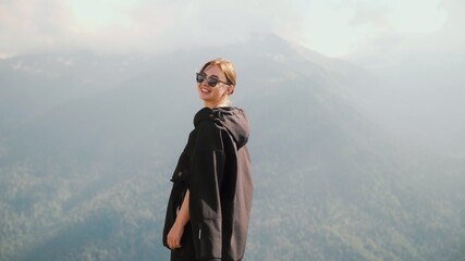 A happy young girl in sunglasses and a black hoodie stands against the background of mountains and smiles