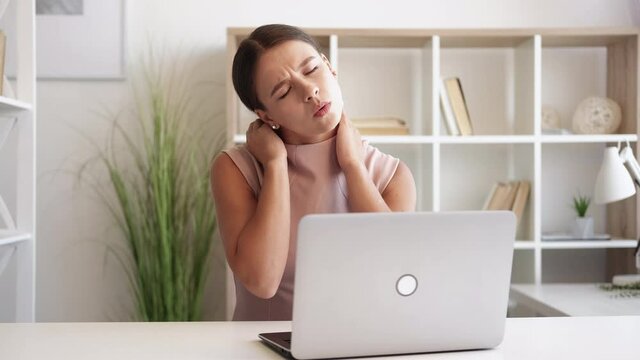 Tired woman. Distance work. Office workout. Exhausted elegant lady stretching neck after typing on laptop sitting light room interior.