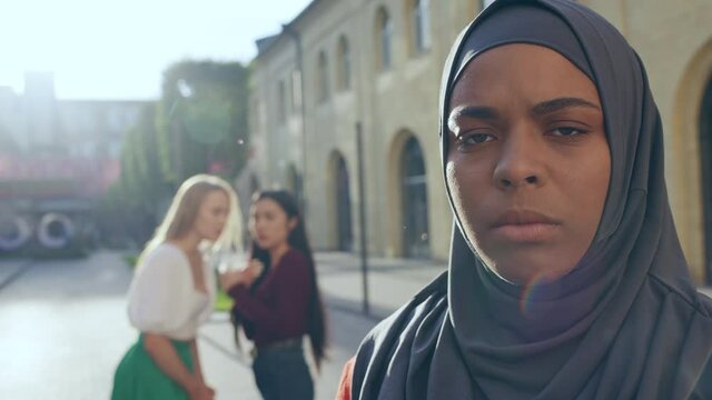 Sad muslim woman looking at camera, gossiping women on background, stereotypes
