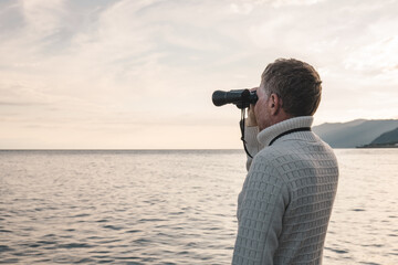 Portrait of a handsome middle-aged man in a white sweater looking through binoculars at the sea in the evening at sunset. Space for text.
