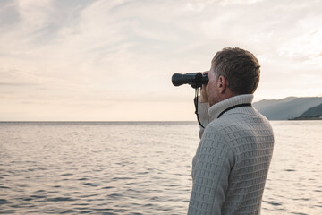 Portrait of a handsome middle-aged man in a white sweater looking through binoculars at the sea in...