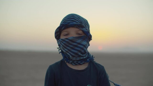 Portrait of 5 years old boy wearing blue checkered keffiyeh standing in desert at dawn. Desert sunrise with tourist kid looking at camera.
