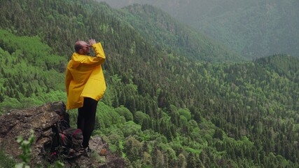 Fototapeta na wymiar A young man in a yellow raincoat stands on a rock in a mountainous area and looks into the distance. Travel and tourism
