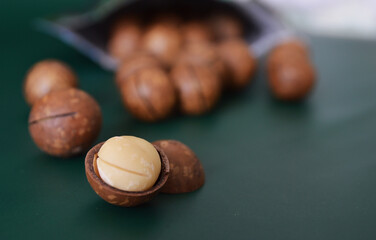 Cracked open macadamia nut in a dark green background of roasted ones rolling out of the bag - obrazy, fototapety, plakaty