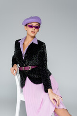trendy young woman in beret, sunglasses and blazer with animal print posing while sitting on chair isolated on grey