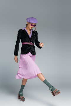 Full Length Of Stylish Young Woman In Beret, Purple Skirt And Blazer With Animal Print Walking On Grey