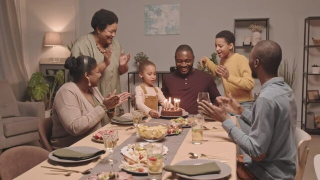 Medium long of two Black girls bringing out and putting birthday cake down on table in front of young happy father, who then blowing out candles and hugging daughters, family members clapping hands