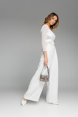 full length of curly young model in white outfit holding purse with zebra print and posing on grey