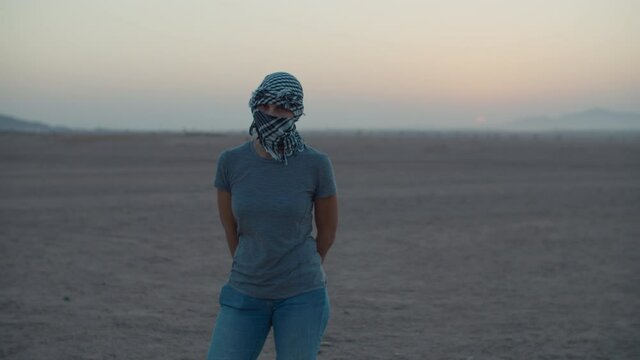 Woman wearing checkered keffiyeh standing in the desert waiting for sunrise looking at camera. Caucasian female tourist standing in desert.