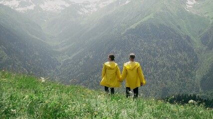 Naklejka na ściany i meble A young man and a woman in yellow raincoats are walking down the slope, holding hands and enjoying the scenery of the mountainous area. Travel and tourism