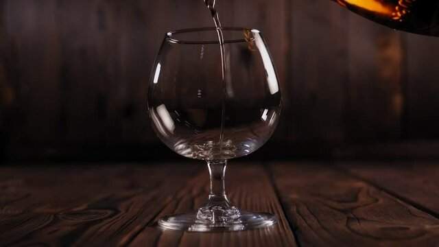 Pouring brandy or cognac from the bottle into the glass against wooden background. Cognac is poured from a bottle into a glass on a wooden background. Slow motion.