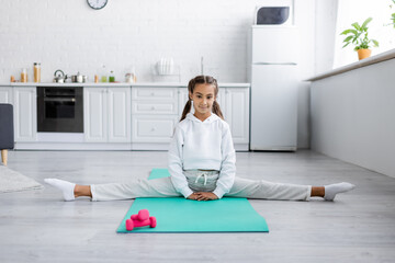 Preteen girl looking at camera while doing split near dumbbells in kitchen
