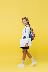 Preteen schoolgirl with backpack and laptop walking on yellow background