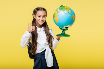 Smiling schoolgirl showing like and holding globe isolated on yellow