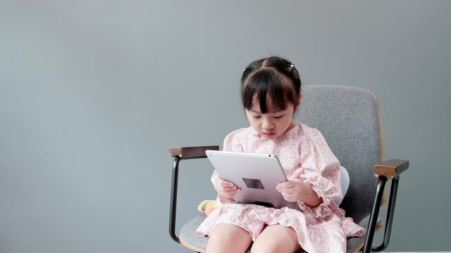 Little asian girl sitting on chair, swing legs, studying online e-learning system on tablet, paying attention to online video. Concept of online learning and social distancing.
