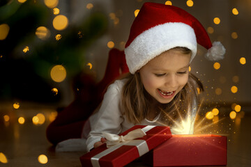 Little girl at dark in red santa hat opens box with magic light and laughs next to christmas tree. Happy child with New Year gift