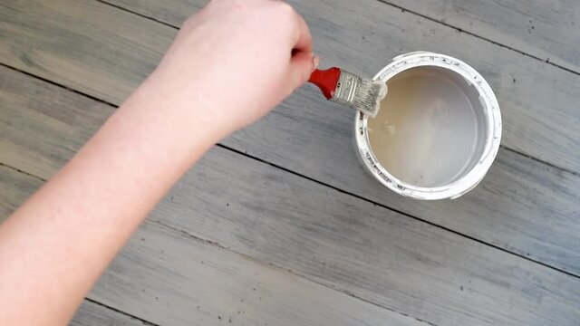 Left-handed woman paints the floor with a brush in grey color. Doing room renovation with light color with lef hand. Brush painting