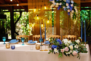 Wedding presidium, banquet table for newlyweds with flowers, greenery, blue and gold color. Lush flower arrangement on the wedding table. Glowing light bulbs. Soft selective focus.