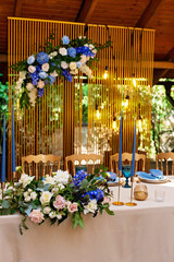 Wedding presidium, banquet table for newlyweds with flowers, greenery, blue and gold color. Lush flower arrangement on the wedding table. Glowing light bulbs. Soft selective focus.
