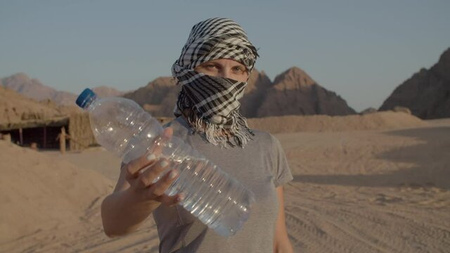 Woman in checkered keffiyeh holding bottle of water standing in the desert. Caucasian female tourist with plastic water bottle. Importance of drinking water.