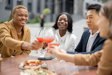 Multiracial business team toasting with wine while celebrating at outdoor cafe. Concept of teambuilding and corporate event. Idea of rest and leisure on job. People sitting at table with pizza