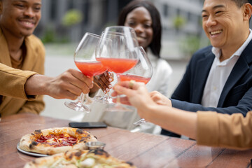 Multiracial business team toasting with wine while celebrating at outdoor cafe. Concept of teambuilding and corporate event. Idea of rest and leisure on job. People sitting at table with pizza