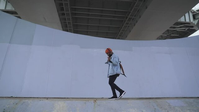 Modern devices to listen music. Joyful young African-American man spbas student wearing denim jacket with headphones dances walking past concrete fence