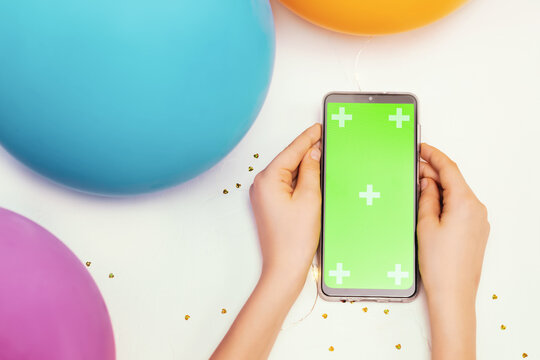 Kid Holiday Banner With Gadget With Green Chroma Key Screen. Child Hands Holding The Smartphone With Green Screen On A White Background With Holiday Decor And Balloons. Mockup For Mobile Apps