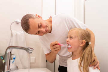 Obraz na płótnie Canvas dad cleans daughter's teeth with a toothbrush in the bathroom, girl smiling