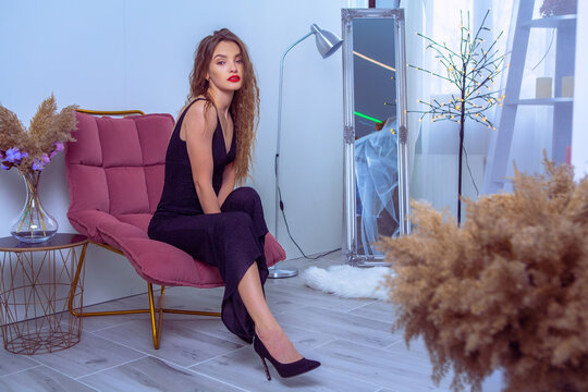 Glamour Woman With Red Lips In Black Overalls Sits On The Chair And Looking At The Camera