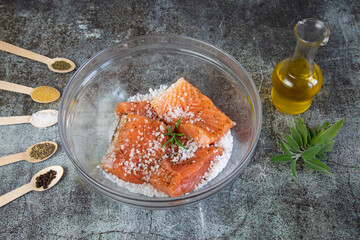  Salting salmon at home,  fillet in a transparent bowl olive oil and various spices in wooden spoons on the table